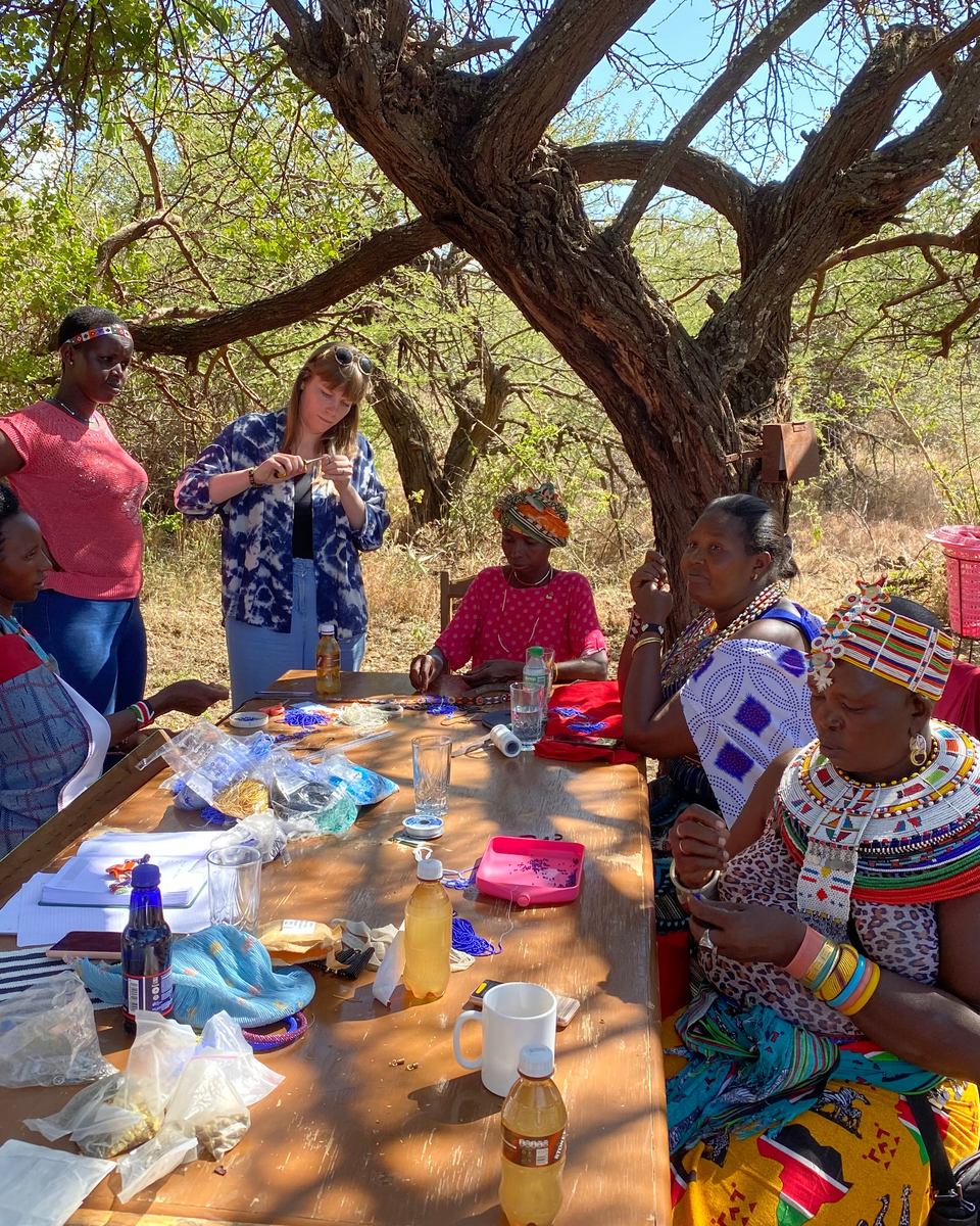 Samburu artisans gathered around a table outdoors under a large acacia tree, engaged in conversation and crafting new jewelry designs.