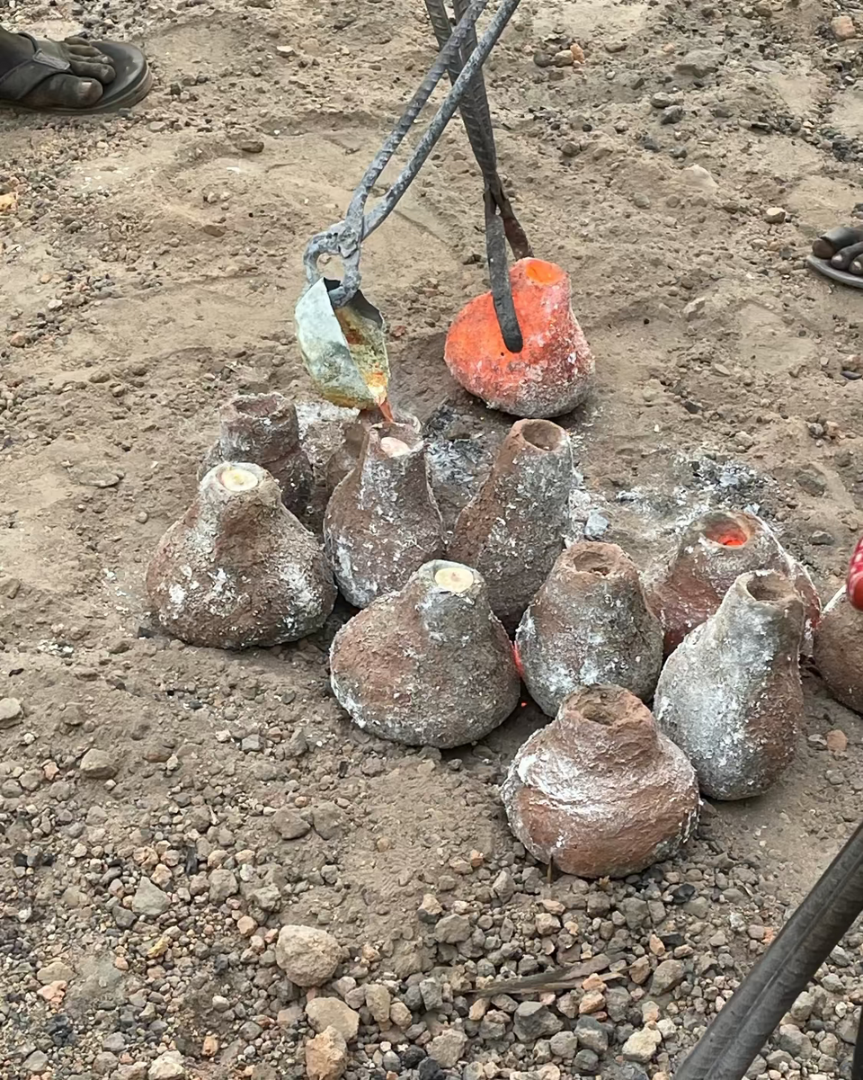 Several rough, handcrafted clay pots with wide flared rims sit on sandy, gravelly ground. Metal tongs holding glowing orange and green molten material are positioned above the pots, suggesting a pottery-making or kiln scene.