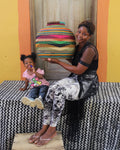 A woman and a young child sit on a black-and-white patterned bench in front of a wooden door, with the woman holding a large, colorful hand-woven basket. The background features a bright yellow wall with orange side panels.