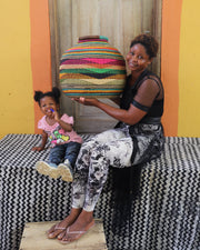 A woman and a young child sit on a black-and-white patterned bench in front of a wooden door, with the woman holding a large, colorful hand-woven basket. The background features a bright yellow wall with orange side panels.