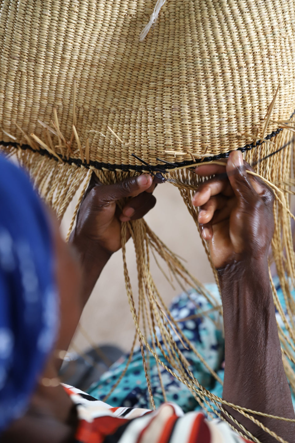 Close-up of a person weaving a large natural straw hat, with their hands holding a thin black cord along the brim's edge. The woven straw has a warm tan color, and the person's hands are visible in the foreground.