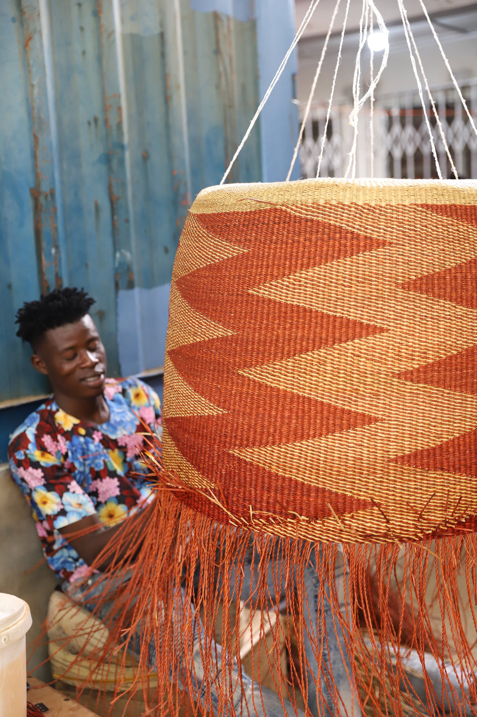 A large woven cylindrical basket in terracotta orange and natural beige chevron pattern and fringe, hanging in a workshop next to a smiling woman.
