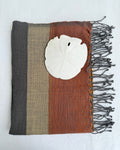 A square woven hand towel with vertical stripes in dark gray, beige, and rust brown, finished with dark fringe on one side and a white shell placed on top, shown against a white background.