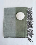 A fhand towel with dark fringes, shown on a white fabric background. The fabric has a two-tone design with sage green and light gray sections, separated by a thin dark stripe, and features a white shell placed on top.