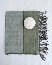 A fhand towel with dark fringes, shown on a white fabric background. The fabric has a two-tone design with sage green and light gray sections, separated by a thin dark stripe, and features a white shell placed on top.
