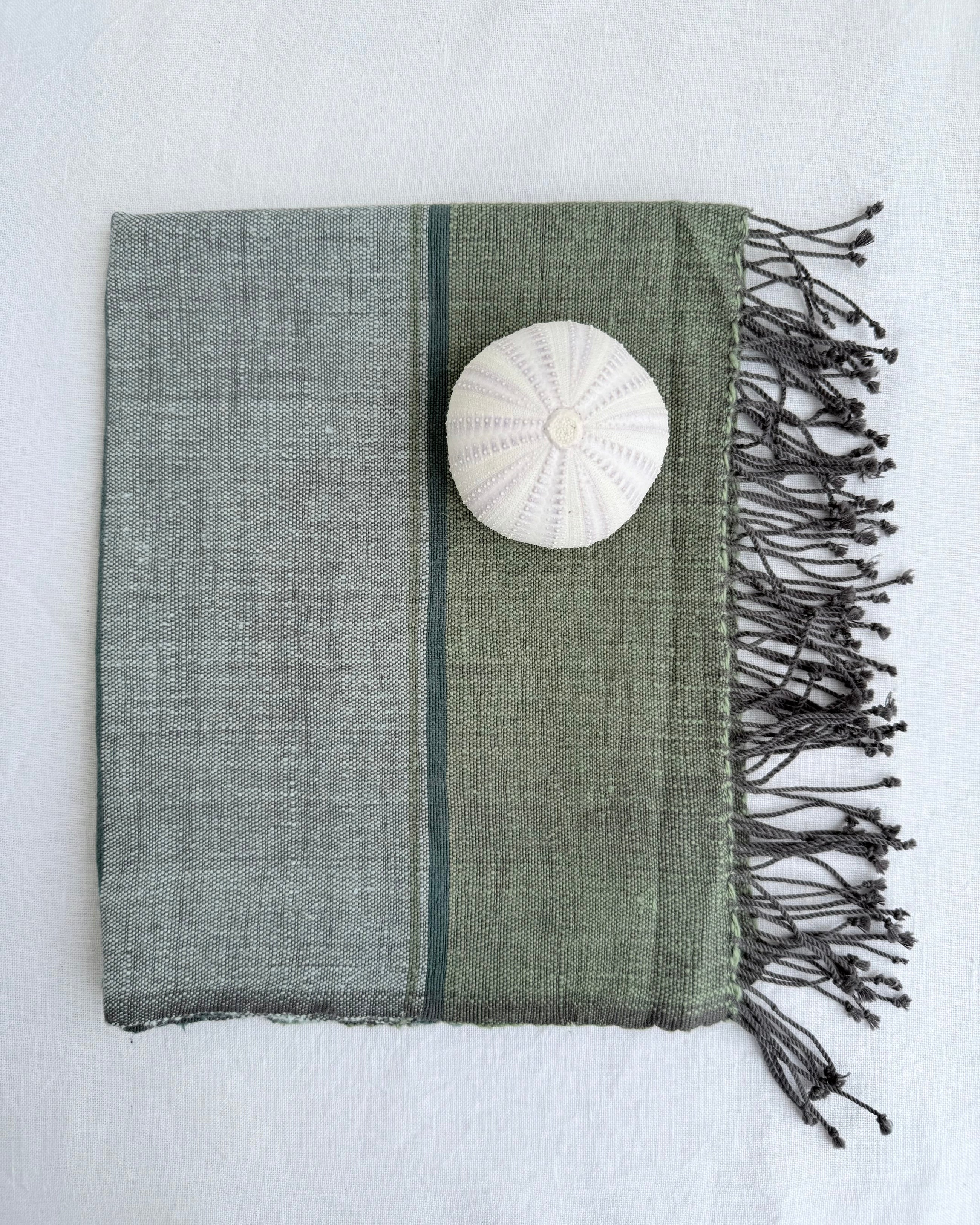 A fhand towel with dark fringes, shown on a white fabric background. The fabric has a two-tone design with sage green and light gray sections, separated by a thin dark stripe, and features a white shell placed on top.