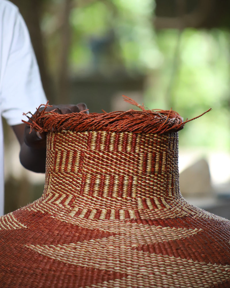 A close-up of a hand-woven basket with a wide, rounded body and a narrow neck, decorated with geometric patterns in reddish-brown and beige. A person's arm and white shirt are visible on the left side, with a blurred outdoor background of green foliage.