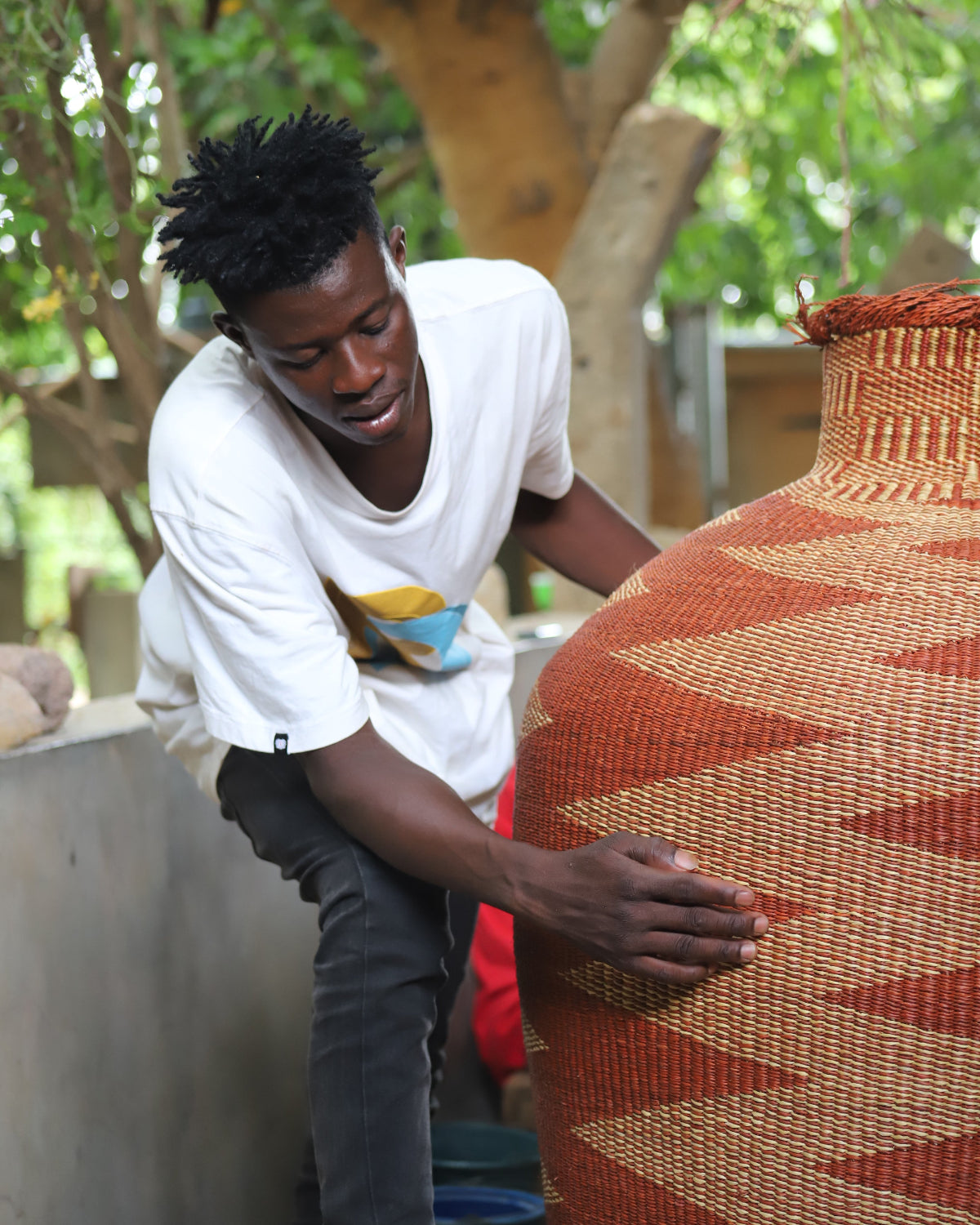 A man in a white graphic t-shirt and dark pants is leaning down to touch a large, hand-woven terracotta and brown basket vase outdoors next to a tree.