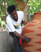 A man in a white graphic t-shirt and dark pants is leaning down to touch a large, hand-woven terracotta and brown basket vase outdoors next to a tree.