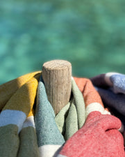 A close-up of a stack of colorful woven towels or blankets draped around a small wooden post, with a blurred turquoise water background.