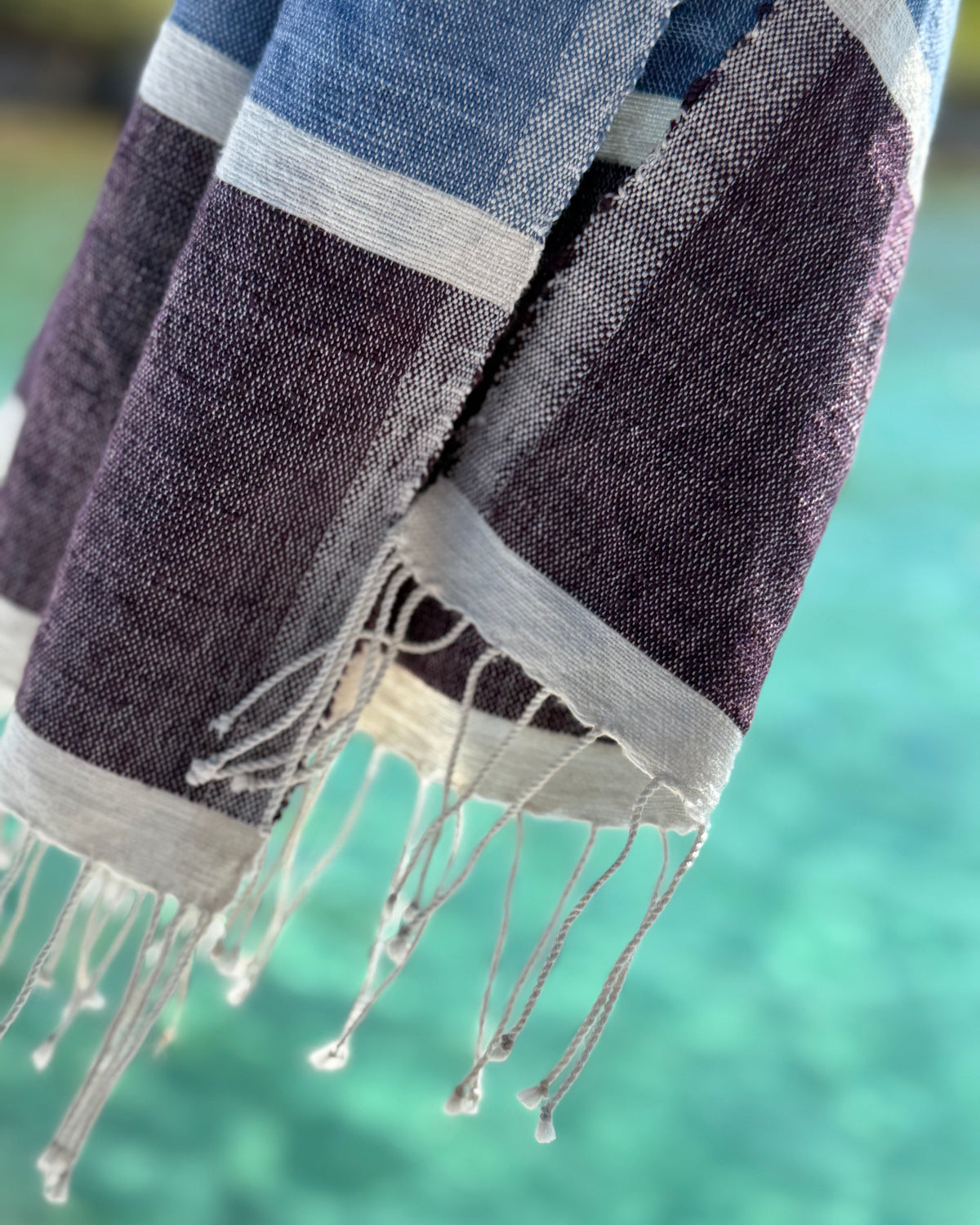 A close-up of a woven Ethjiopian-style towel or scarf with fringed edges, featuring horizontal stripes of dark purple, slate blue, and light gray, set against a blurred turquoise background.