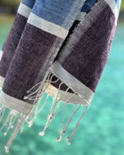 A close-up of a woven Ethjiopian-style towel or scarf with fringed edges, featuring horizontal stripes of dark purple, slate blue, and light gray, set against a blurred turquoise background.