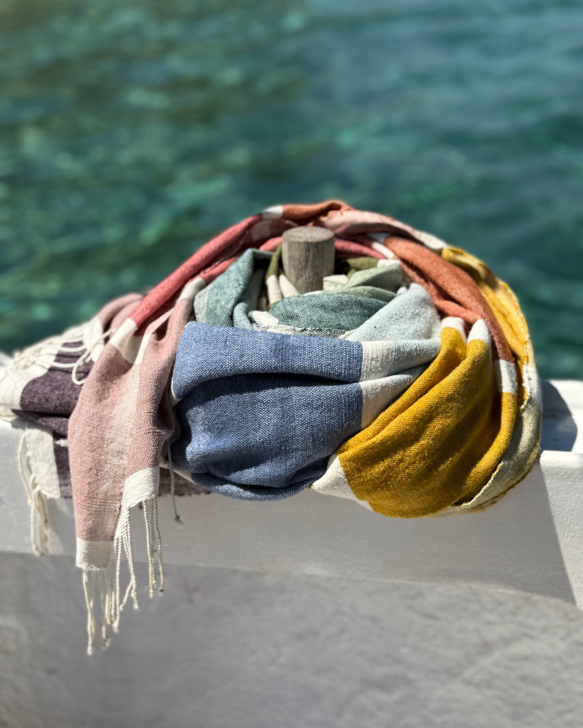 A folded stack of colorful woven throw blankets with fringed edges, draped over a white railing with a blurred blue-green water background.