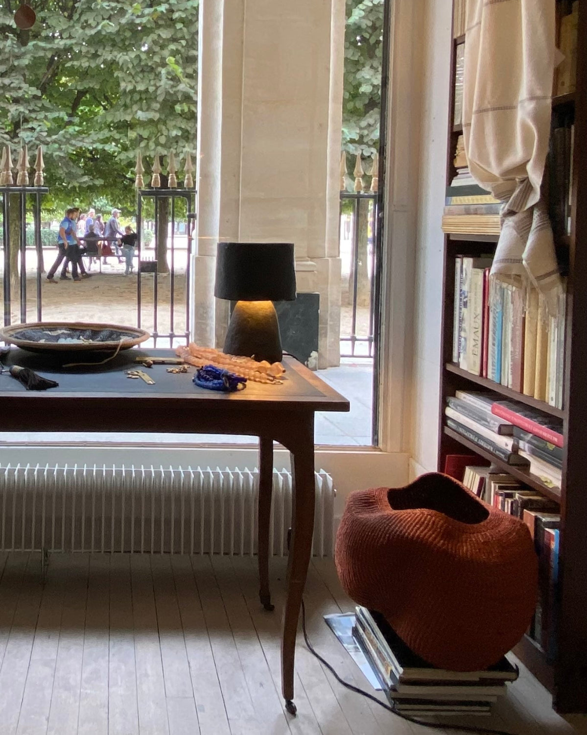 A rustic wooden table with a black lamp and decorative bowl in a cozy room with a large window looking out onto a green garden, next to a bookshelf and a terracotta-colored basket.