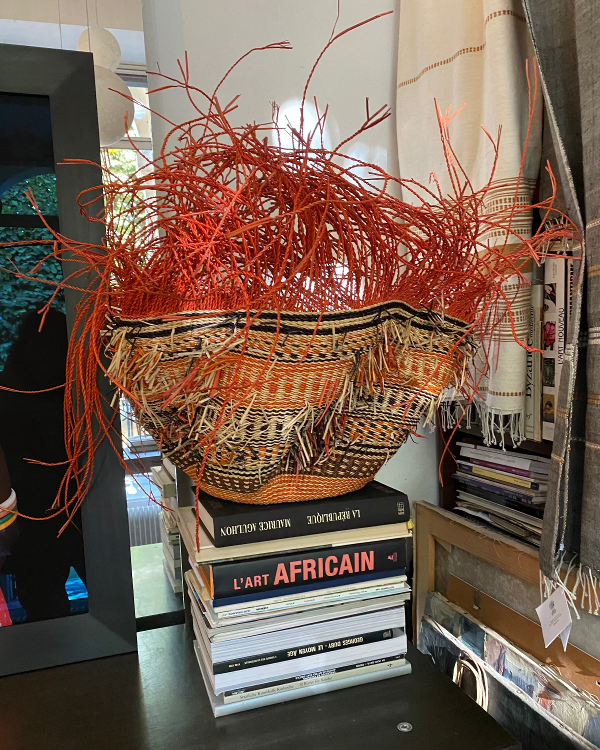 A large decorative woven basket with bright orange dried grass or straw stems overflowing from the top, displayed on a stack of books on a dark table in an indoor room setting.