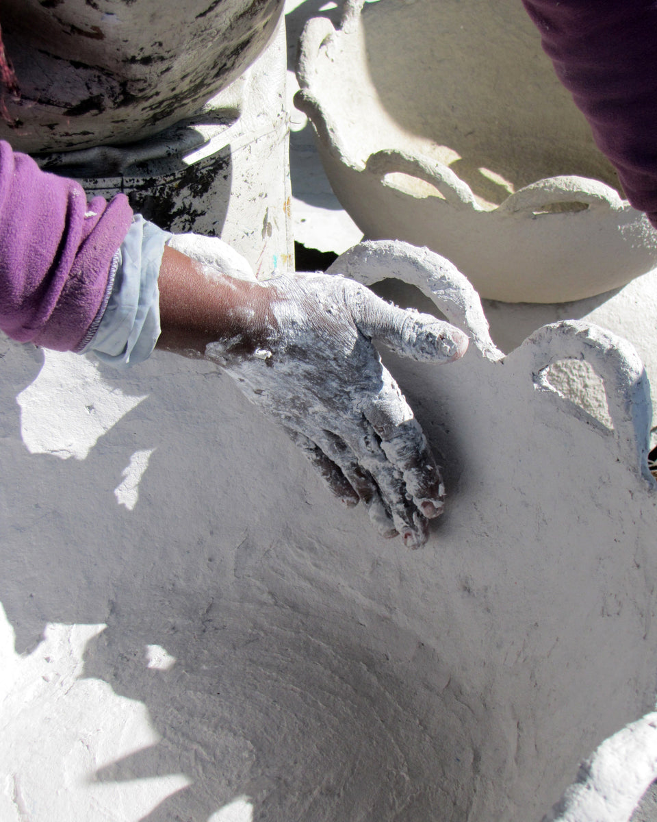 A close-up of a person's hand covered in white plaster or clay, smoothing the inside of a handmade vessel. The person wears a purple sleeve, and other clay pots and bowls are visible in the background.