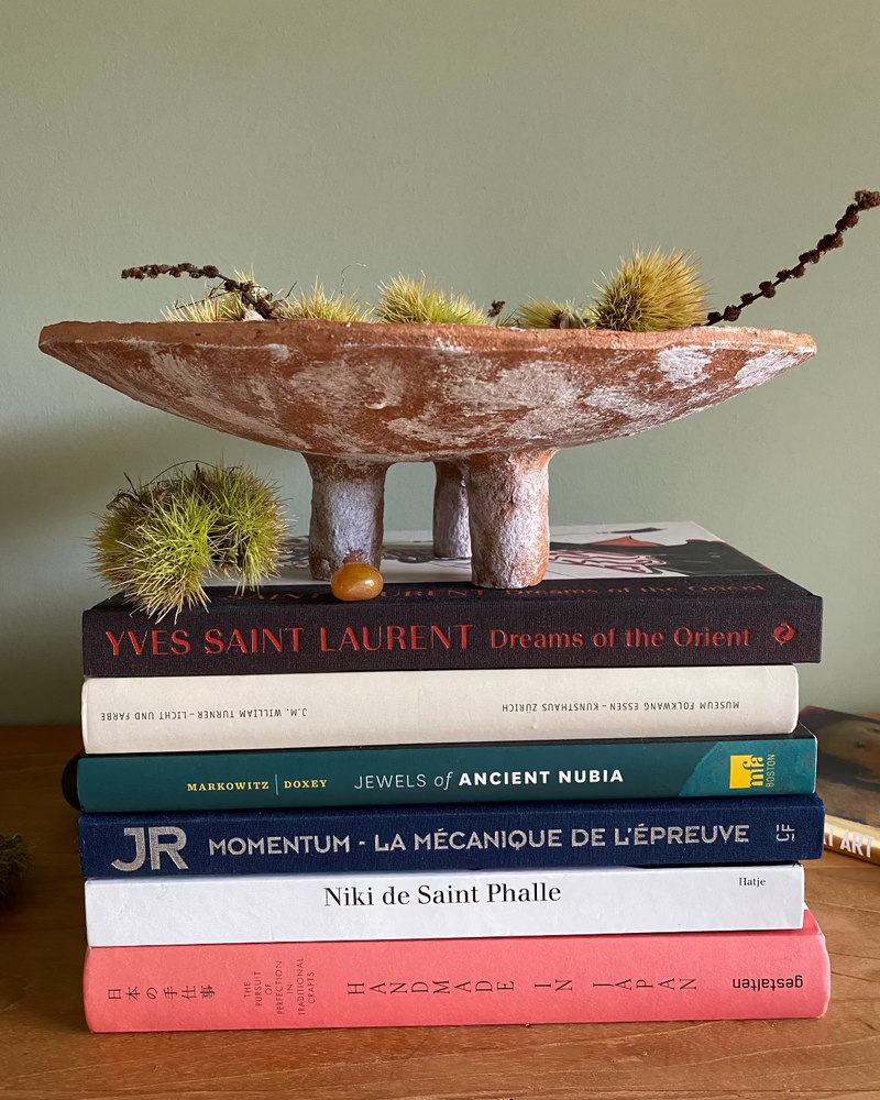 A terracotta-colored ceramic tripod bowl  with a white finish sits on top of a stack of books on a wooden surface. The bowl contains dried green seed pods or grasses, and a few spiky green botanical elements rest beside it.