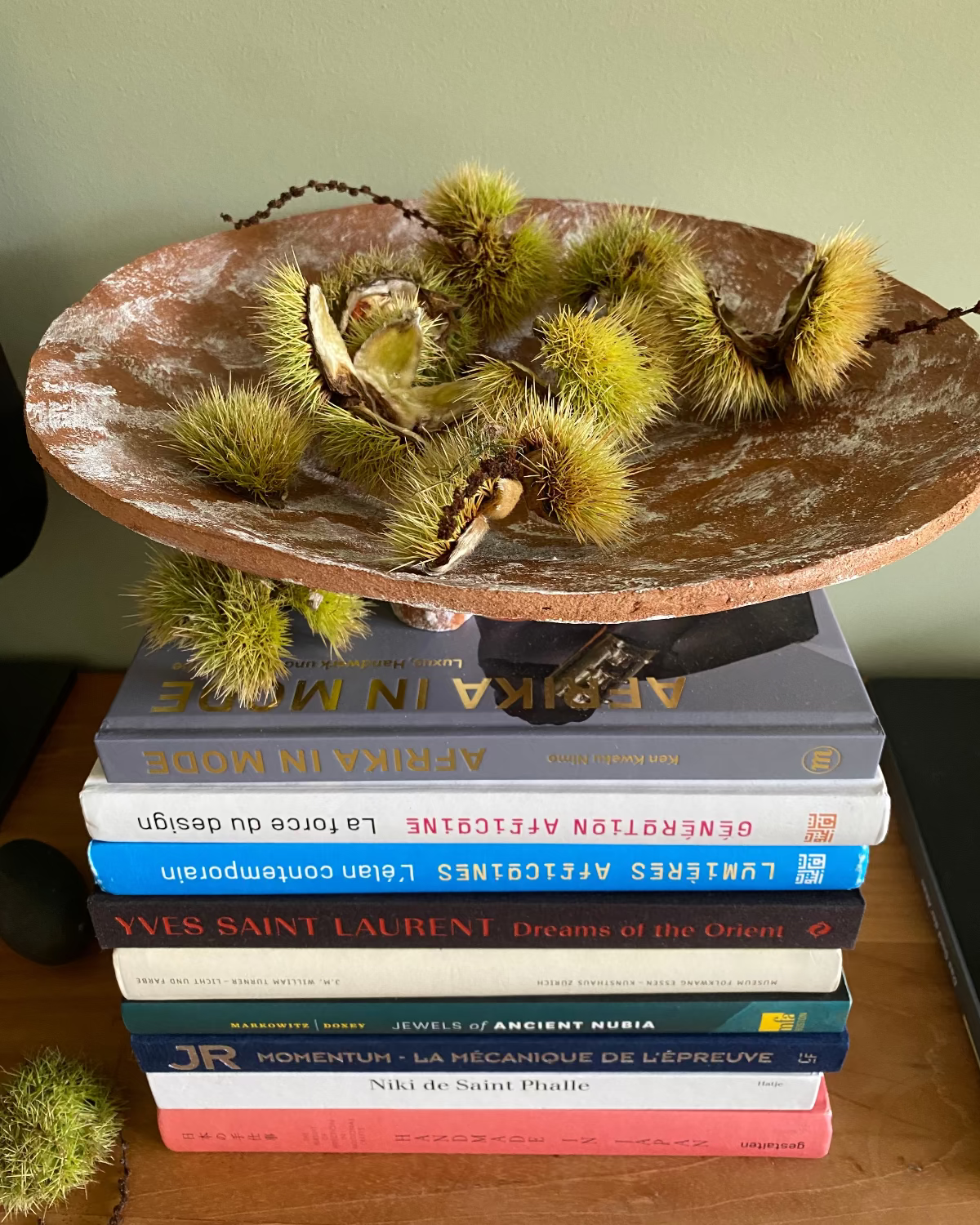 A round ceramic bowl made from paper pulp filled with green chestnuts sits on top of a tall stack of books on a wooden surface against a muted green wall.