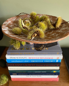 A round ceramic bowl made from paper pulp filled with green chestnuts sits on top of a tall stack of books on a wooden surface against a muted green wall.