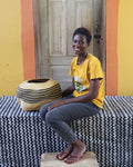 A smiling woman sits on a black-and-white patterned bench in front of a wooden door, wearing a yellow graphic T-shirt and gray pants. A large woven basket sits beside her on the bench.