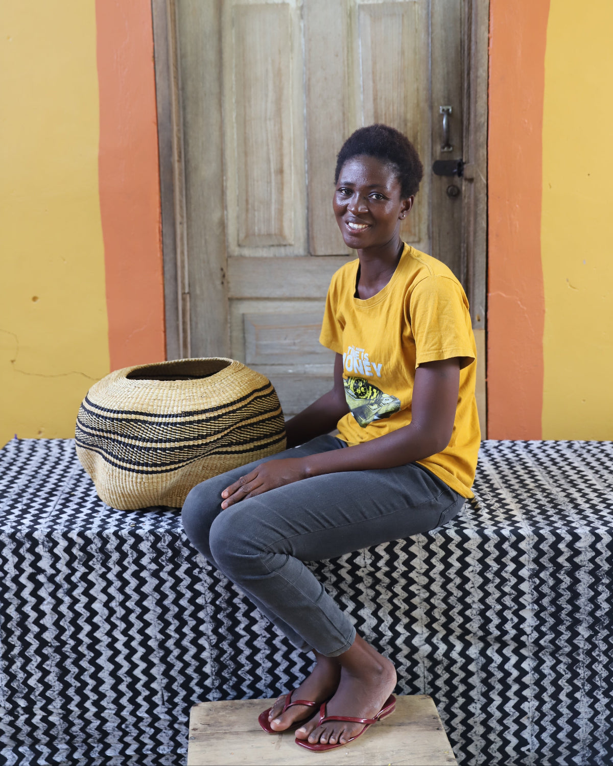 A smiling woman sits on a black-and-white patterned bench in front of a wooden door, wearing a yellow graphic T-shirt and gray pants. A large woven basket sits beside her on the bench.