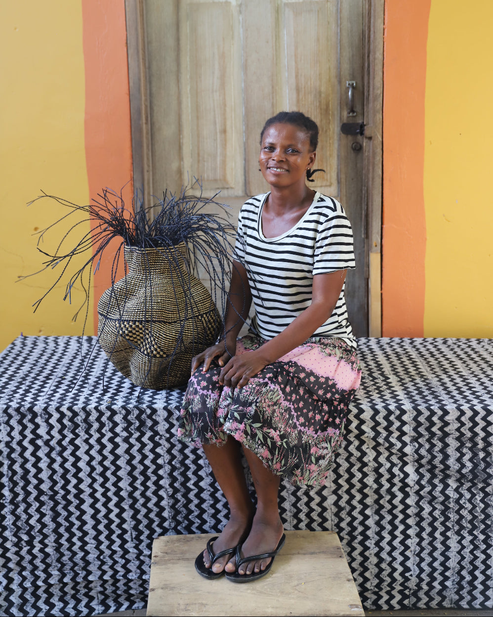 A woman sits on a patterned bench in front of a wooden door, wearing a black and white striped short-sleeve top and a pink and black floral skirt. A woven basket with dried branches sits beside her on the bench.