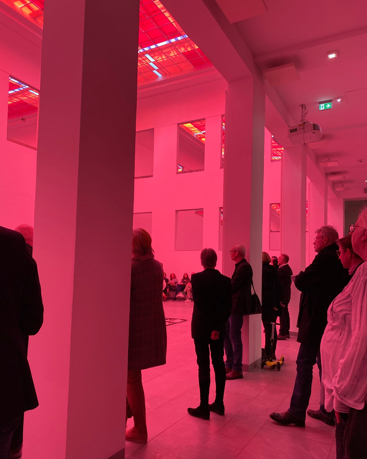 A group of people standing and viewing an art gallery installation with a striking pink and red light projection on the walls and ceiling.