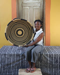A woman sits on a black-and-white patterned bench holding a large round woven basket with a sunburst design. She is wearing a white sleeveless top and gray pants, with a yellow and orange wall and a wooden door behind her.