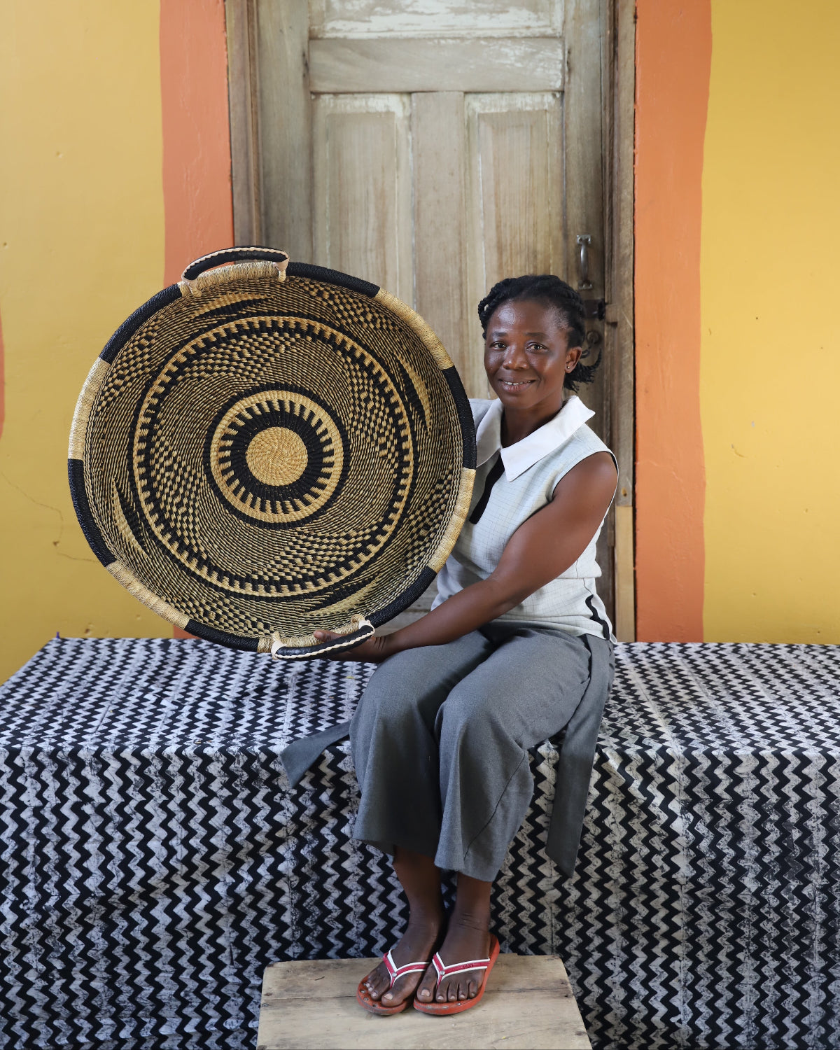 A woman sits on a black-and-white patterned bench holding a large round woven basket with a sunburst design. She is wearing a white sleeveless top and gray pants, with a yellow and orange wall and a wooden door behind her.