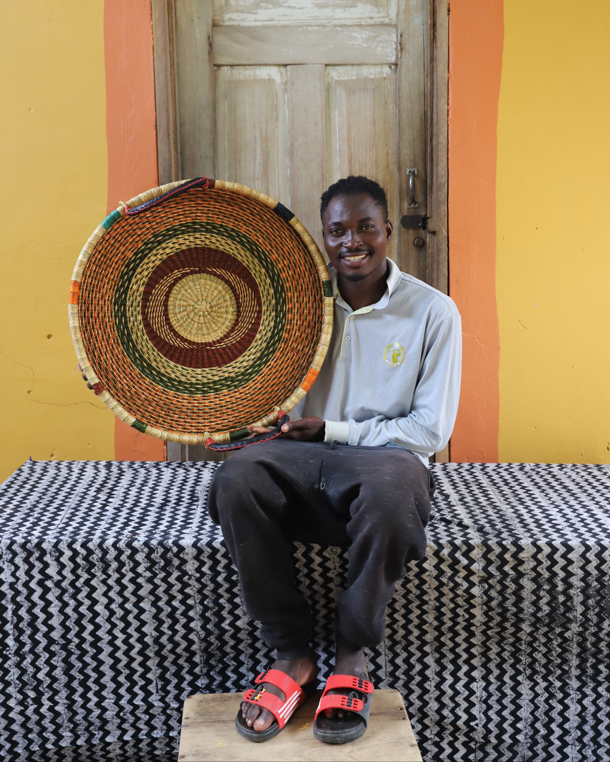 A person sitting on a bench wearing a gray shirt and red sandals, holding up a colorful handwoven wall hanging against a yellow and orange wall.