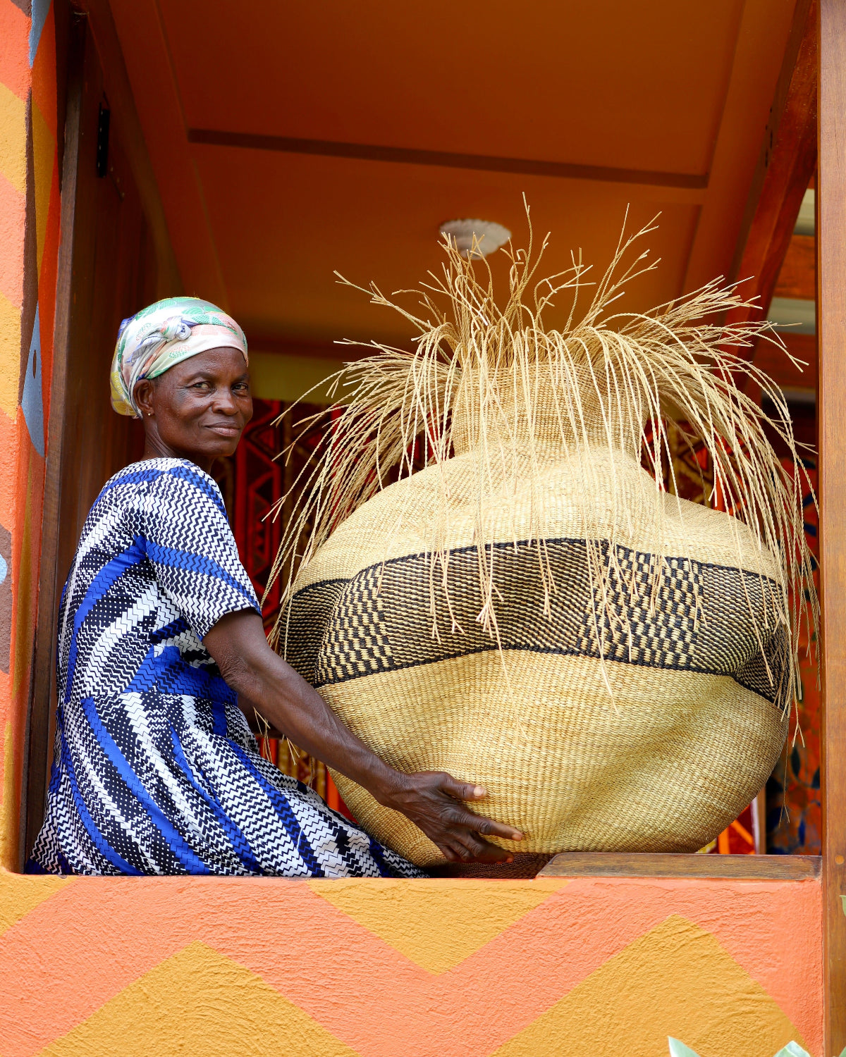 An artisan presenting her large, handcrafted woven basket, resembling a tia, towards a colorful building.