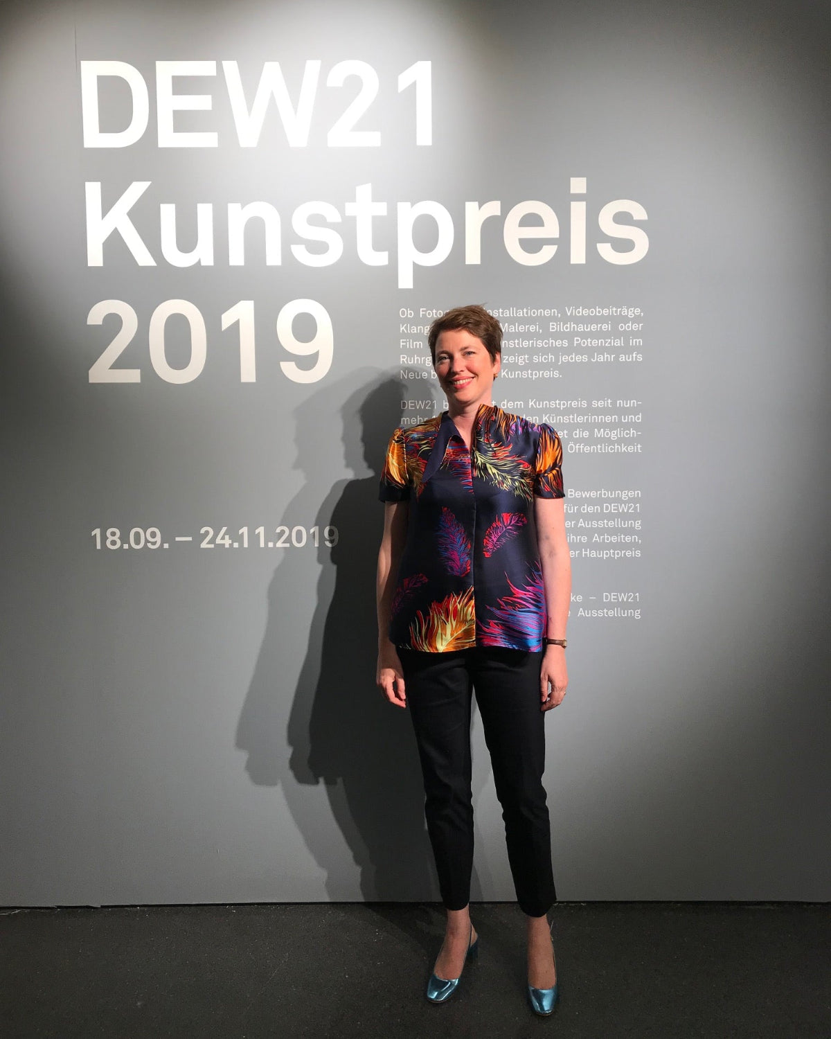 A woman stands in front of a gray wall displaying the text 'DEW21 Kunstpreis 2019' with white lettering. She is wearing a colorful short-sleeve button-up shirt with a vibrant abstract print in orange, yellow, blue, and purple, paired with black pants and blue heels.
