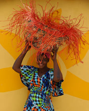 A person holding a colorful basket as headwear with a vibrant red and yellow design, holding the hat up against a yellow background.