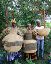Three people standing outdoors in front of a red metal frame and leafy trees, each holding a large woven basket made from natural straw and tan woven panels.