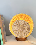 A round woven basket lamp with a spiral pattern, glowing with warm yellow light from within, resting on a dark wooden base on a white surface next to books.
