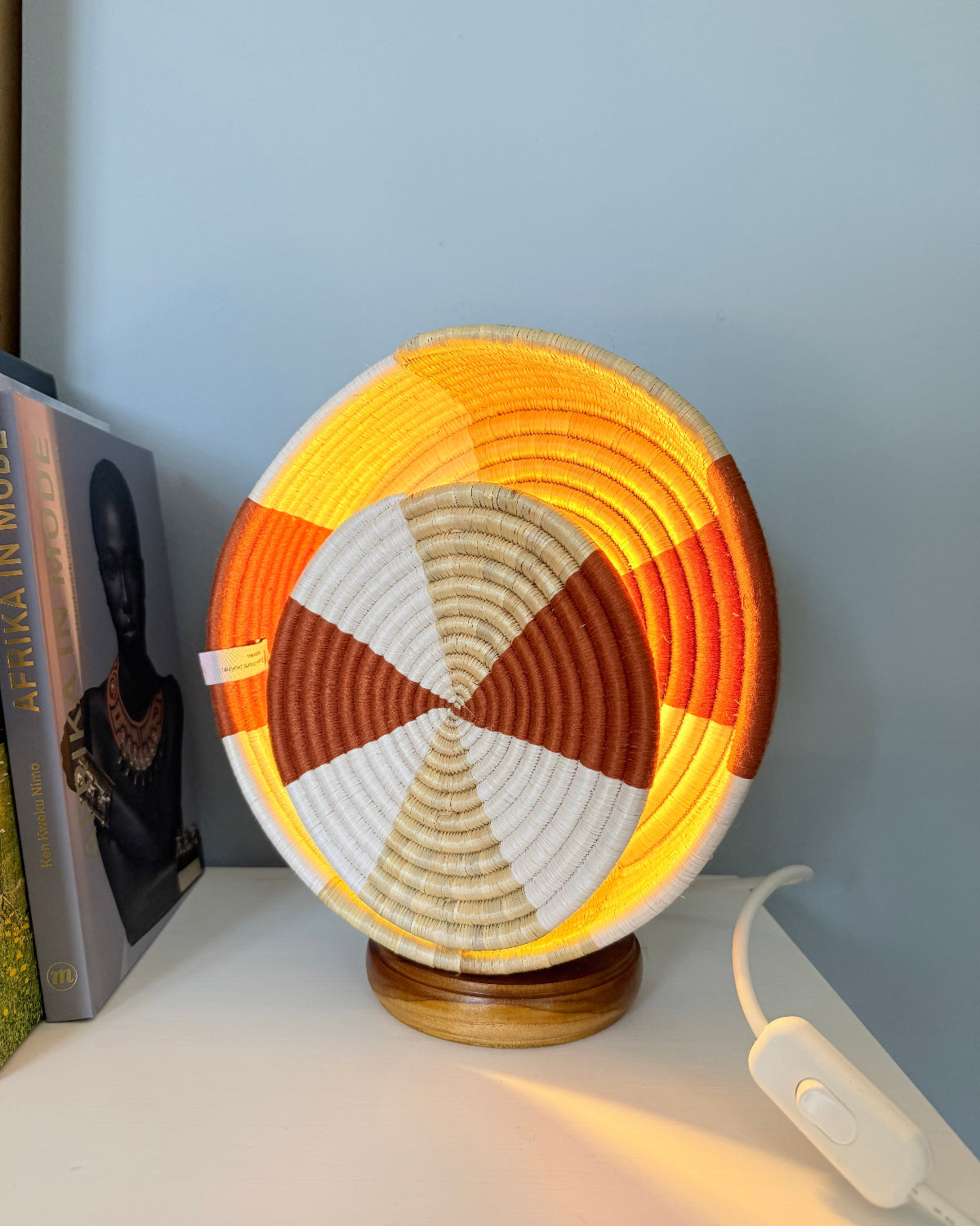 A circular woven basket lamp with a geometric pattern in orange, yellow, brown, and white, illuminated from within to cast a warm glow on a white surface against a light blue wall.