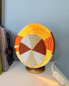 A circular woven basket lamp with a geometric pattern in orange, yellow, brown, and white, illuminated from within to cast a warm glow on a white surface against a light blue wall.