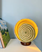 A round woven basket lamp with concentric rings of yellow, green, and beige, sitting on a dark wooden base on a white table against a light blue wall.