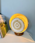 A round woven basket lamp with a spiral pattern in natural beige and white, glowing with a warm yellow light, sitting on a white shelf next to books against a light blue-gray wall.