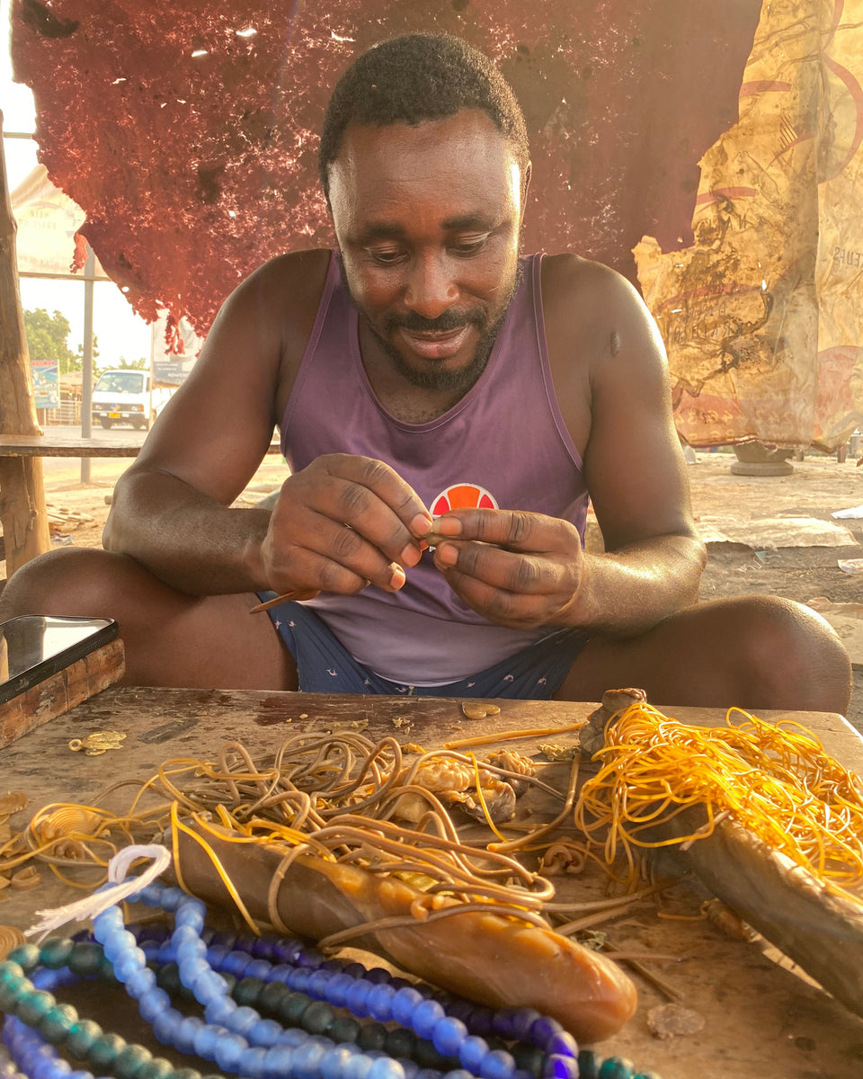 A Ghanaian artisan  crafts a subtle wax jewelry pendant with his hands. The table is covered with wax cords, blue and purple beads, and other jewelry-making materials.