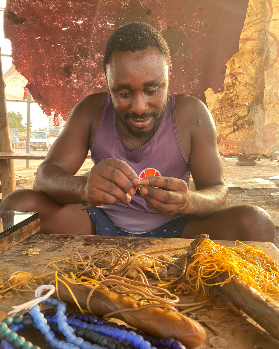A man wearing a purple tank top sits cross-legged at a wooden table, crafting small orange beads with his hands. The table is covered with yellow cords, blue and purple beads, and other jewelry-making materials.