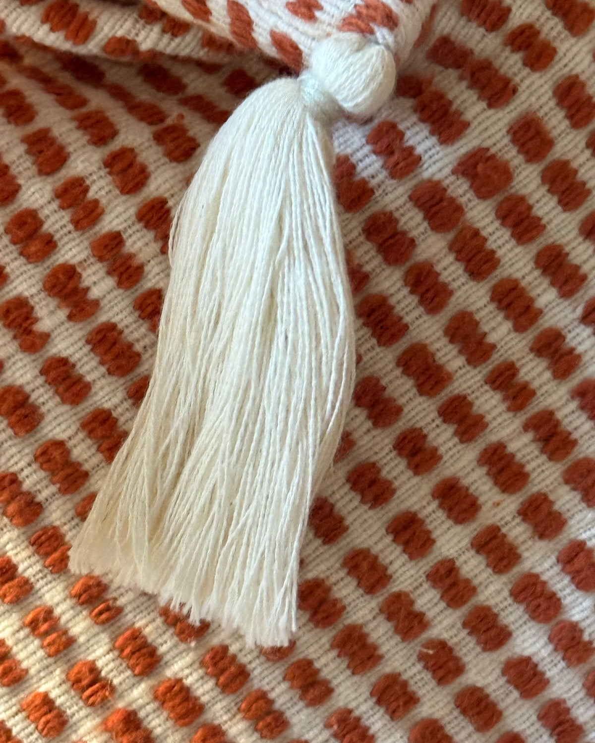 A close-up of a cream-colored tassel tie attached to a textured woven fabric with a rust-orange and cream checkered pattern.