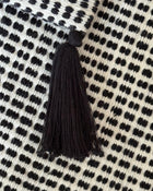A close-up of a black tassel with a round black top, resting on a cream-colored woven textile with a black dotted pattern.