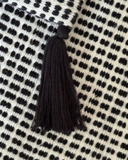 A close-up of a black tassel with a round black top, resting on a cream-colored woven textile with a black dotted pattern.