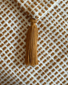 A close-up of a woven textile with a repeating brown dot pattern on an off-white background, featuring a matching brown tassel hanging from the top edge.