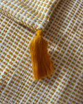 A close-up of a woven textile with a mustard yellow tassel, featuring a white and mustard yellow checkered pattern.
