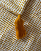 A close-up of a woven textile with a mustard yellow tassel, featuring a white and mustard yellow checkered pattern.