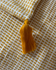 A close-up of a woven textile with a mustard yellow tassel, featuring a white and mustard yellow checkered pattern.