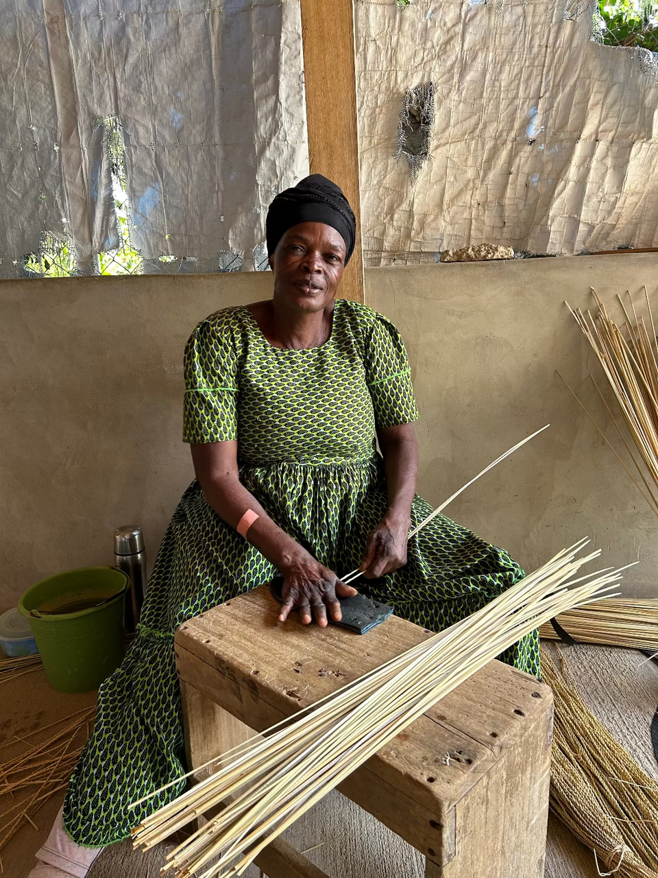A woman wearing a green and black patterned dress and black headwrap sits on a wooden stool, holding a long thin reed while processing bundles of similar reeds in a rustic workshop setting.