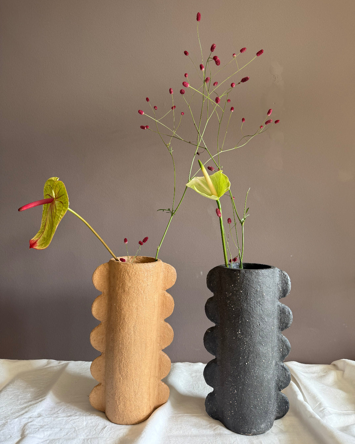 Two sculptural ceramic vases with scalloped edges, one in warm beige and one in dark charcoal gray, styled with floral stems against a taupe wall and white fabric surface.